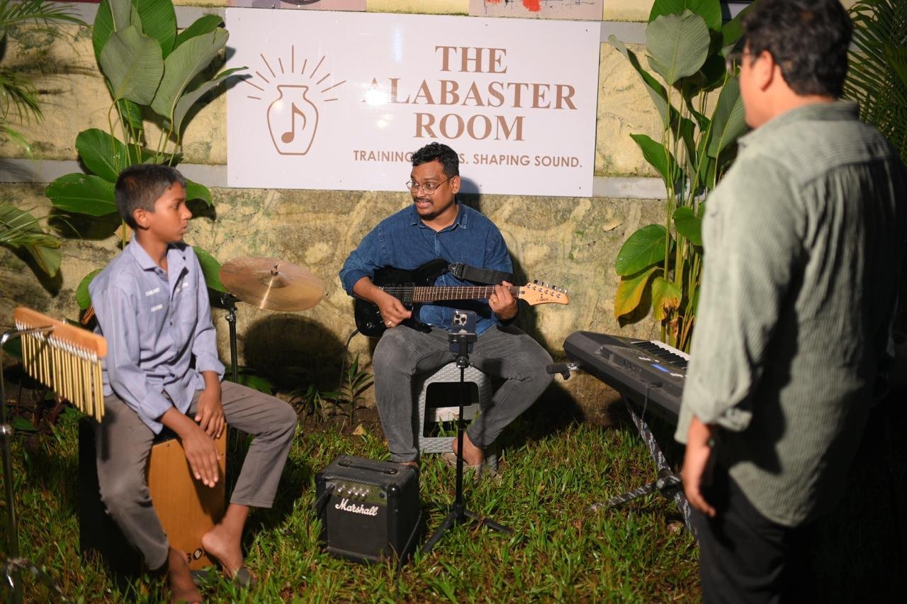 A young student on cajon beside a guitarist under The Alabaster Room sign